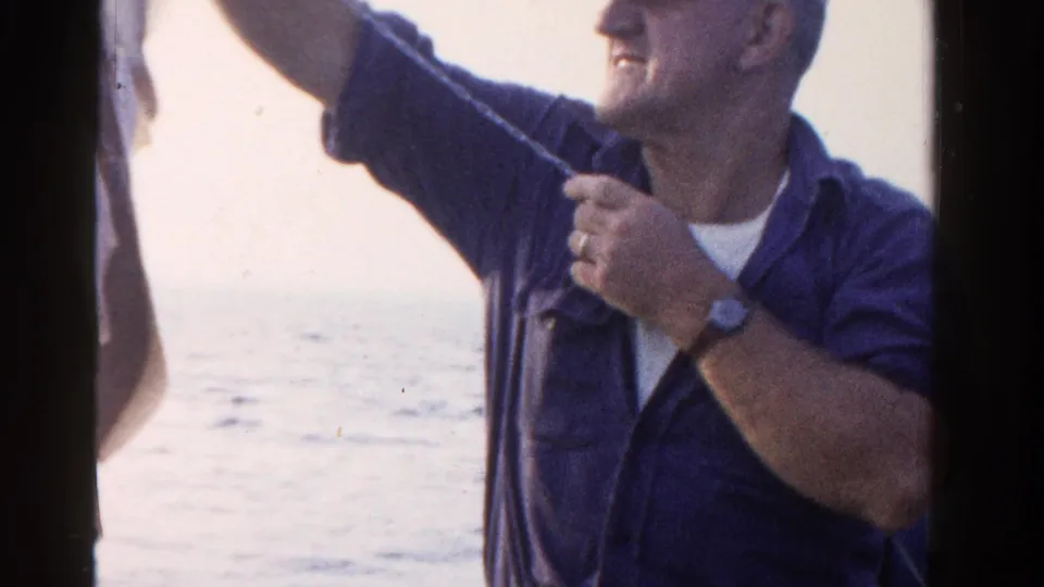 A man in a blue shirt smiles and raises his arm beside open water in this 1958 Ontario home-movie frame.