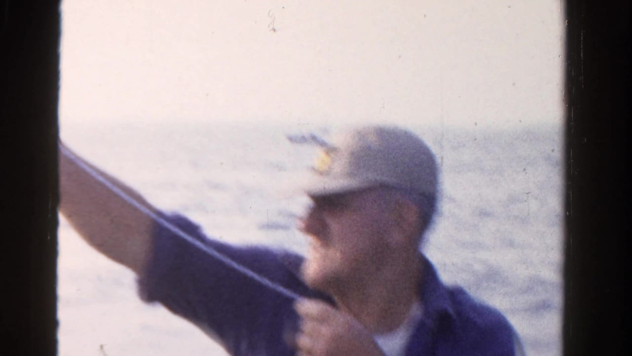 Man in a cap on a boat, partly blurred, holding fishing line near open water.