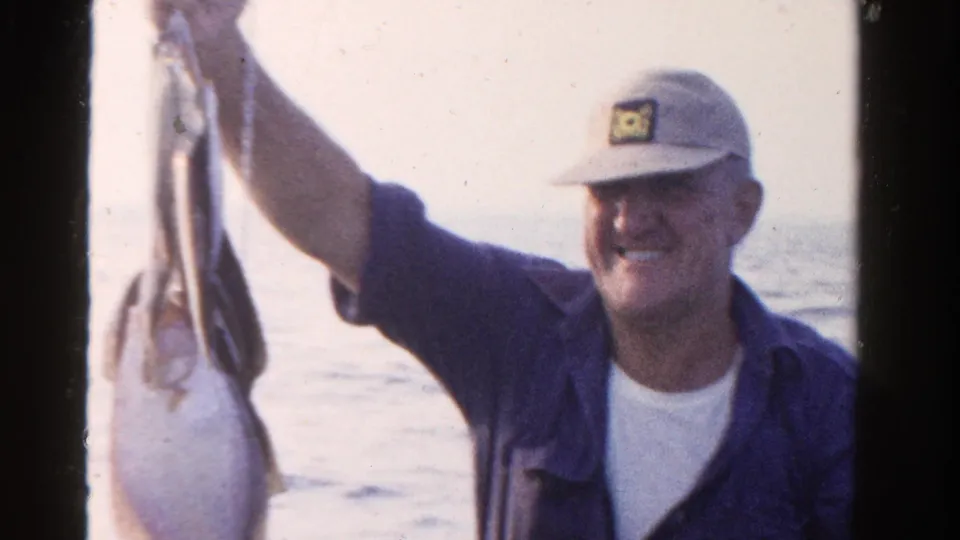 A smiling man in a cap holds up his fish catch on a boat in 1958 Ontario home-movie footage.