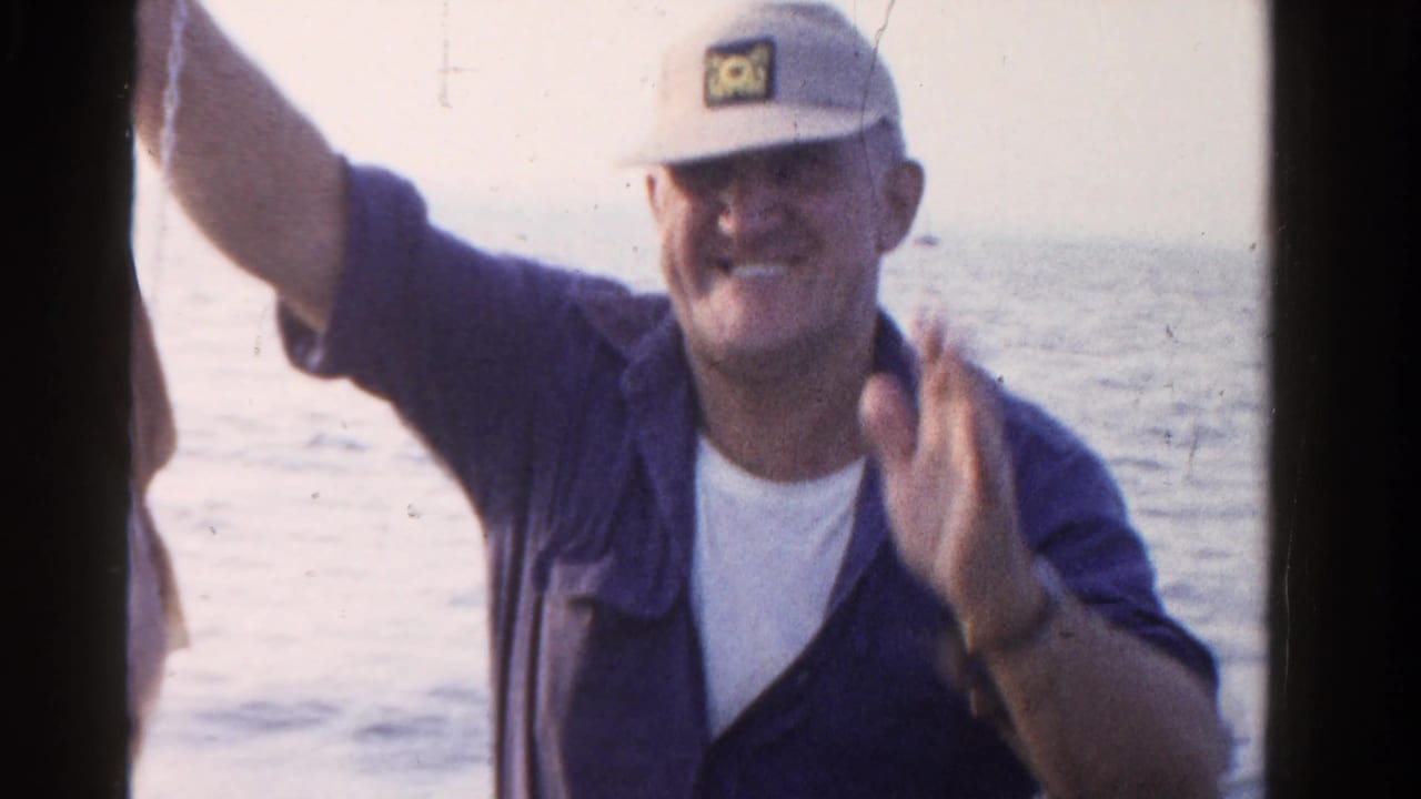 In 1958 Ontario home-movie footage, an adult man in a cap and blue shirt sits in a boat with open water behind him.