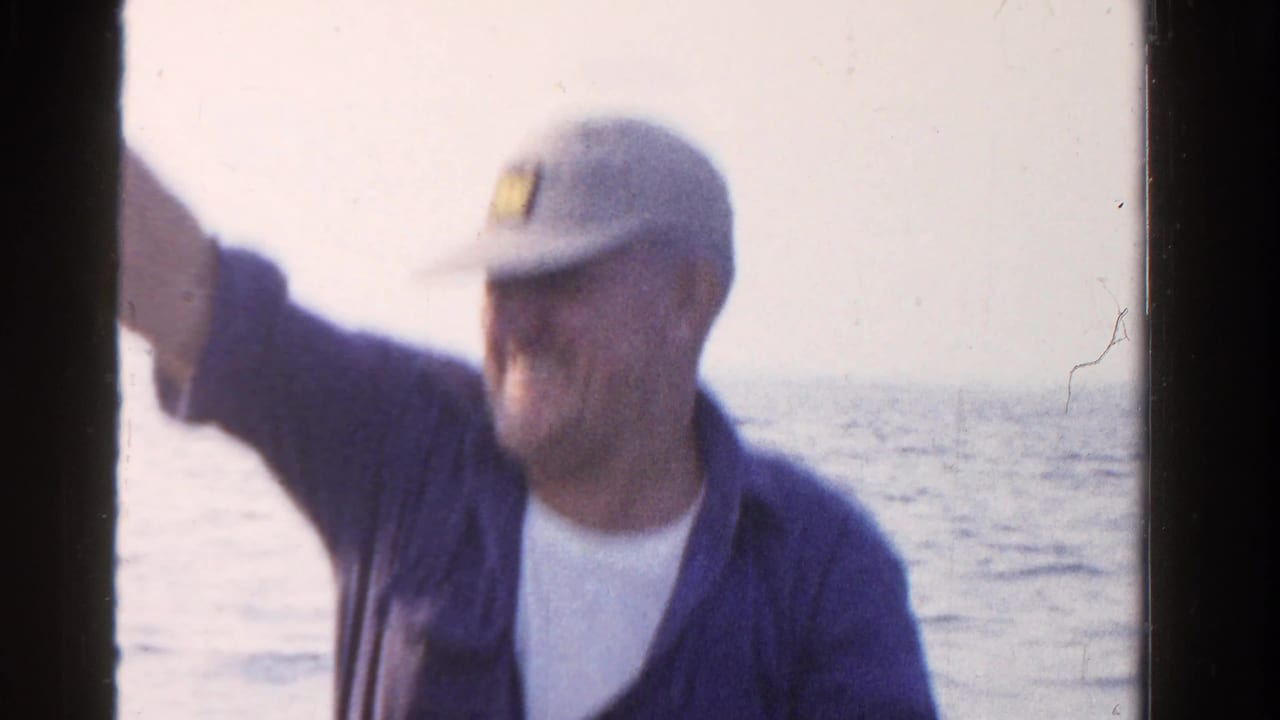 The seated man gestures toward the camera from a boat, with Ontario open water visible in the 1958 home-movie frame.