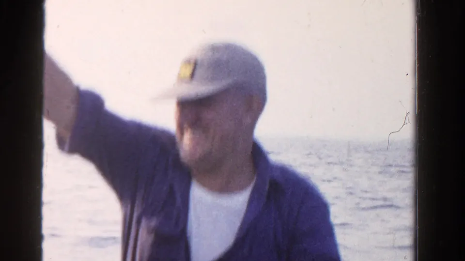 The seated man gestures toward the camera from a boat, with Ontario open water visible in the 1958 home-movie frame.
