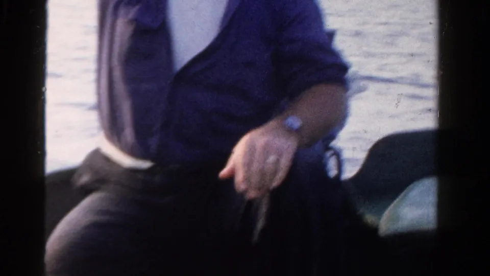 A close 8mm frame shows the man seated in the boat, his wristwatch and clothing visible against the water backdrop.