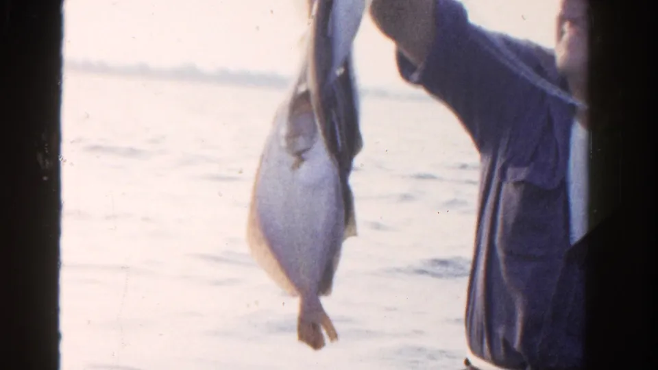 The fisherman stands at the right edge of the frame, raising his catch against bright water and a faint shoreline.