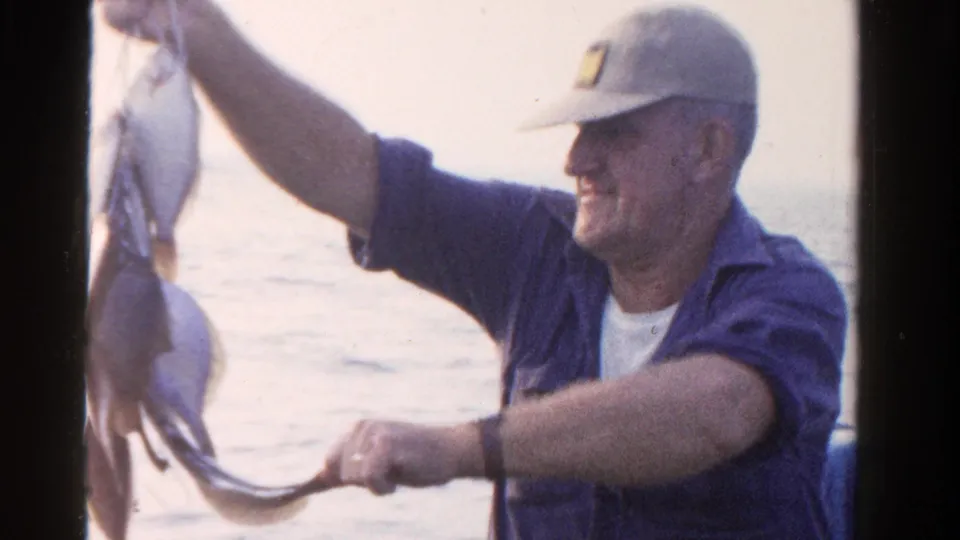 A smiling fisherman in a light cap and blue shirt holds up multiple fish from a boat in supported 1958 Ontario footage.