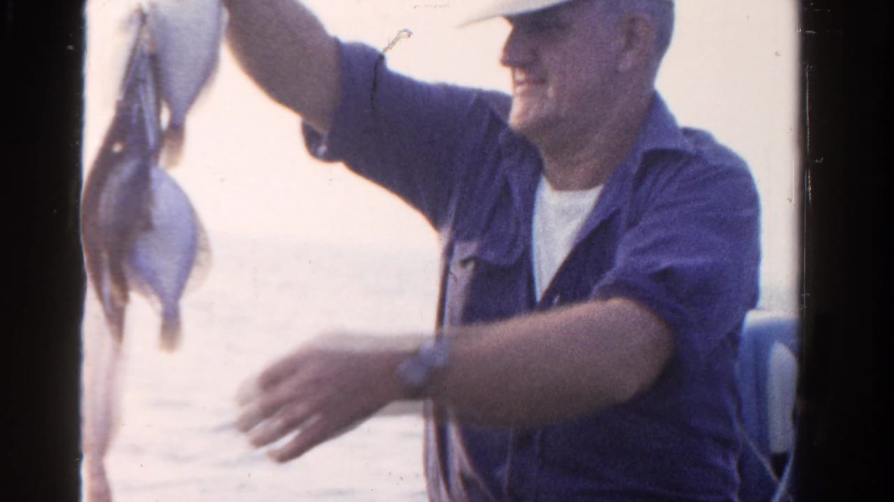 In Ontario, Canada in 1958, a man in a blue shirt and cap raises a string of caught fish while standing on a boat.