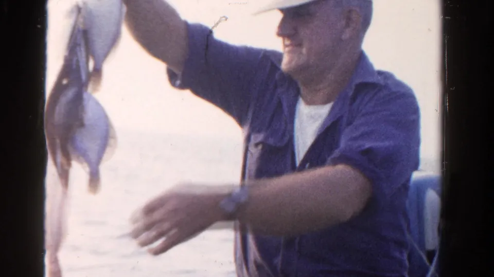 In Ontario, Canada in 1958, a man in a blue shirt and cap raises a string of caught fish while standing on a boat.
