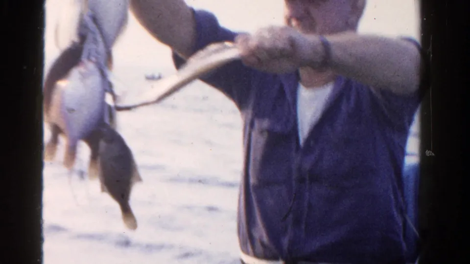 A 1958 Ontario home-movie frame shows a man displaying a line of fish, with water and a partly visible person in the background.