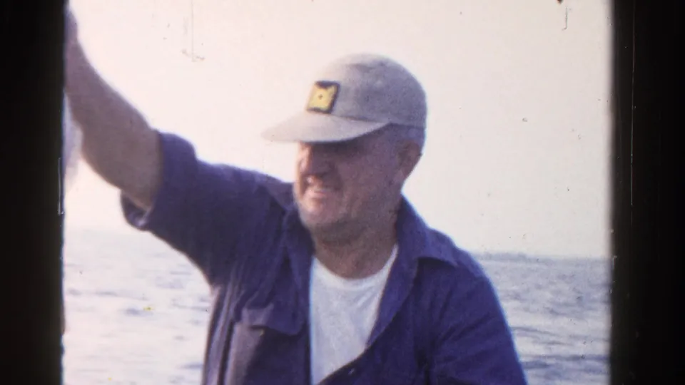 A smiling man in a blue shirt and cap raises his arm on a boat, with open water visible in this 1958 Ontario home-movie frame.