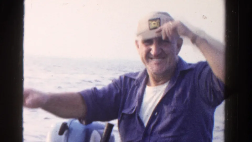 Smiling older man in a small Ontario boat adjusts or touches his cap while facing the 1958 home-movie camera.