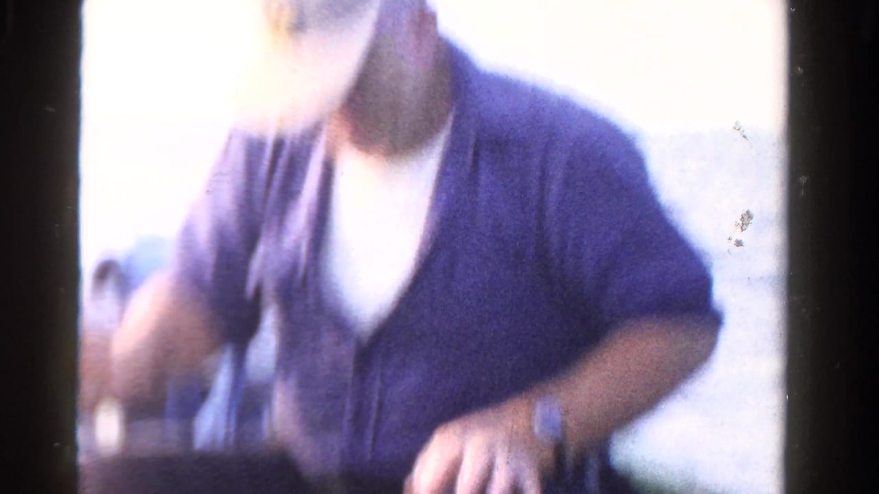 Adult man in a cap and purple shirt seated in a small boat on a lake.