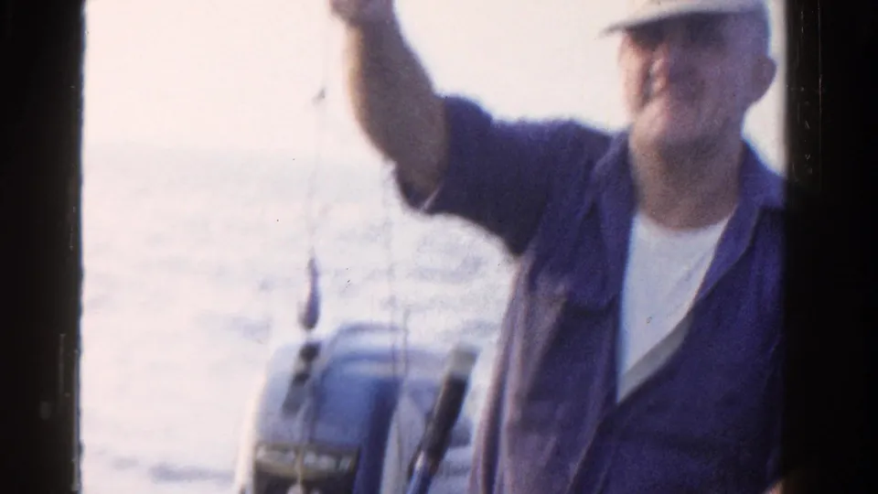 The fisherman raises one arm in the boat, with lake water and a small motor visible in this Ontario home-movie frame.