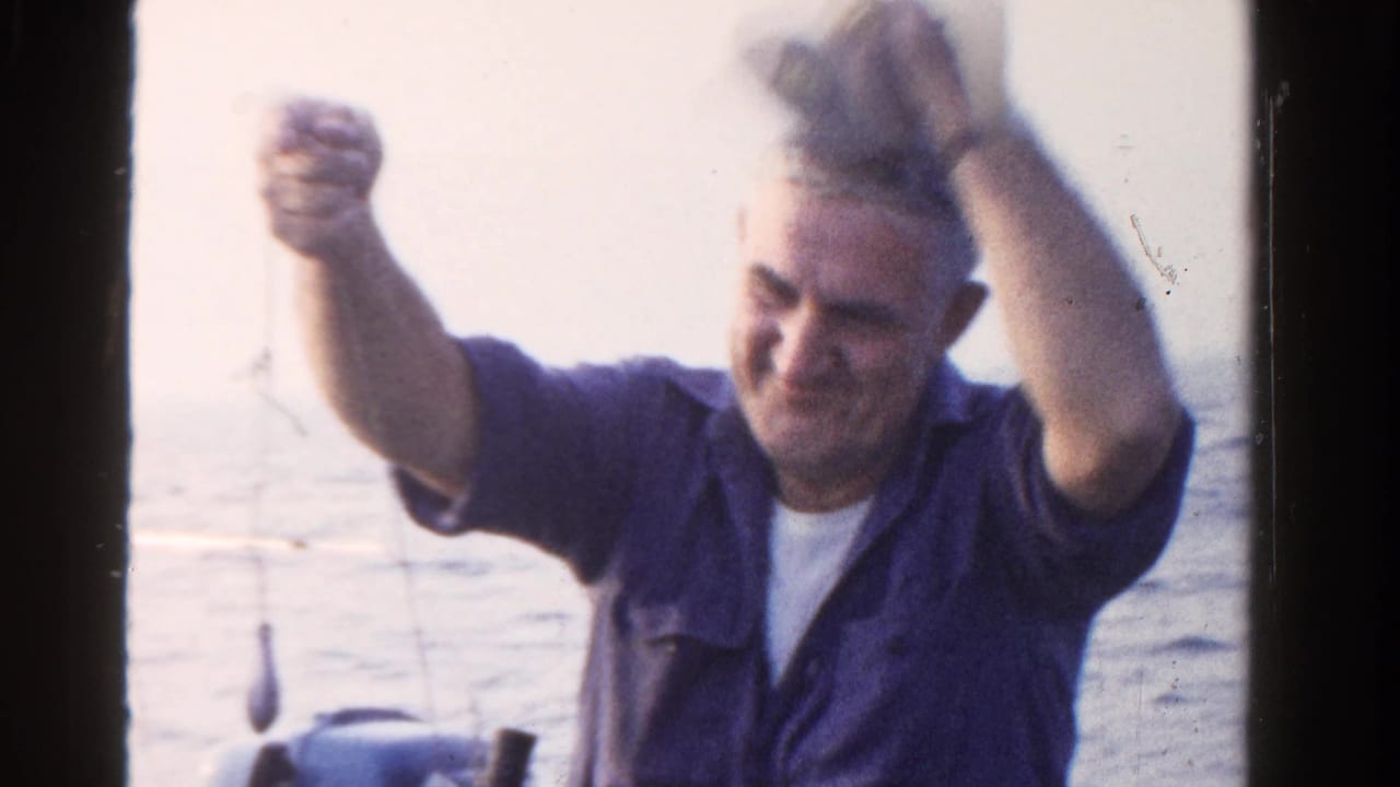 In 1958 Ontario home-movie footage, a man in a blue shirt and white cap lifts his hands while seated on a boat by calm water.