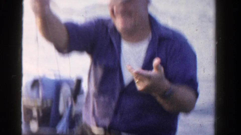 A seated adult man in a blue shirt holds fishing line on a boat, with Ontario water and shoreline in the background.