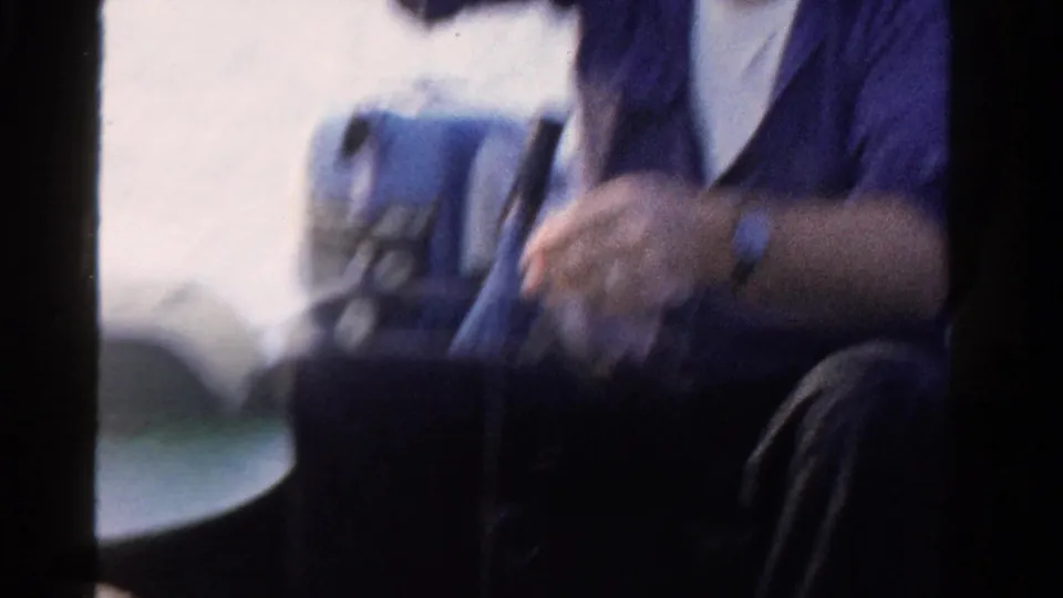A soft 8mm frame shows the seated man in a blue shirt moving on the boat, with water and boat equipment partly visible.