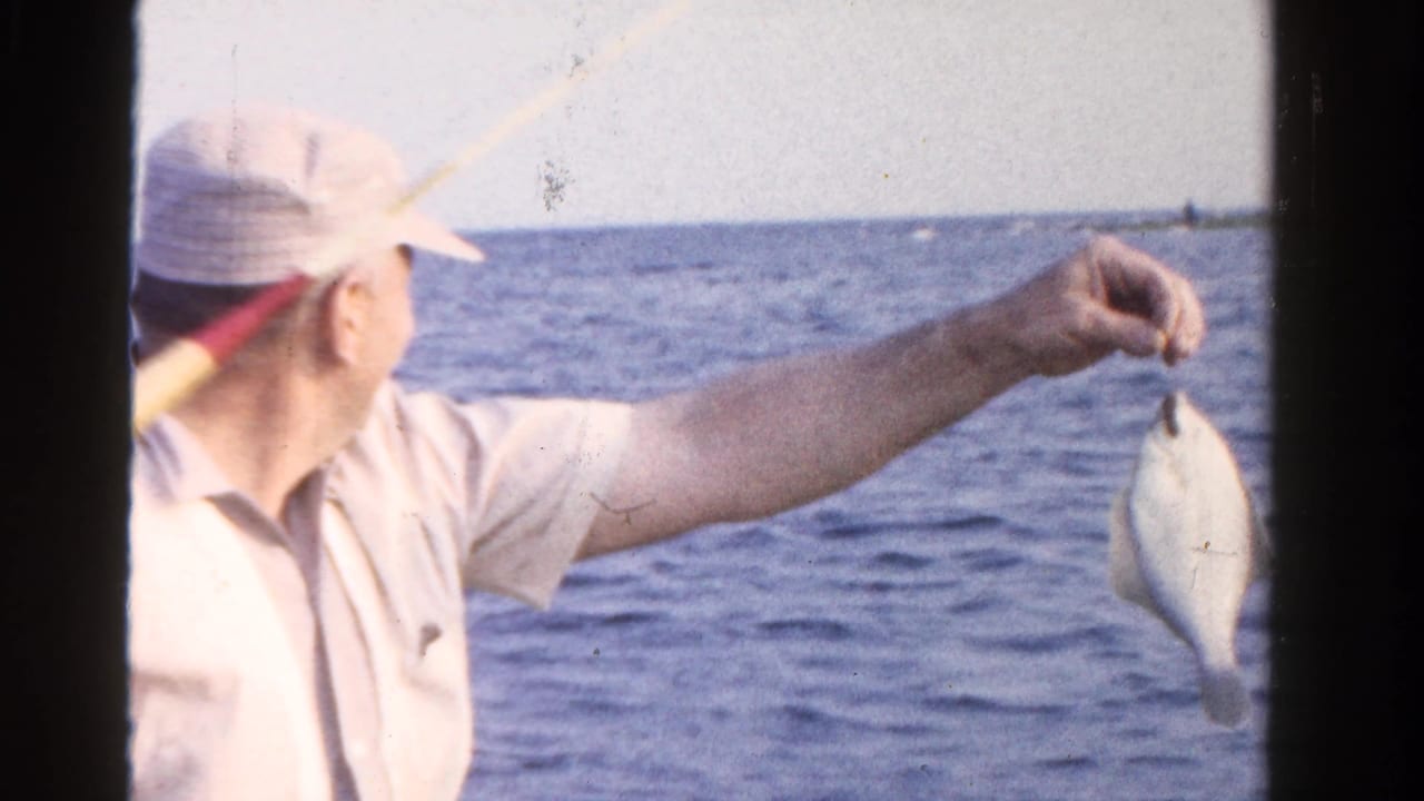 Older man in a pale cap holds a small fish over open water from a boat.