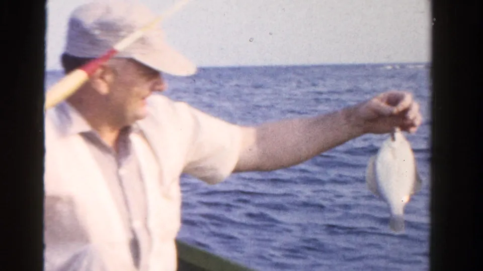 A smiling older man in 1958 Ontario footage holds a small fish outward from a boat with open water behind him.