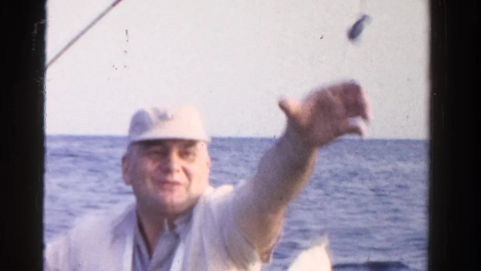 An adult man in a pale cap gestures toward the camera while aboard a small boat on Ontario water in 1958.