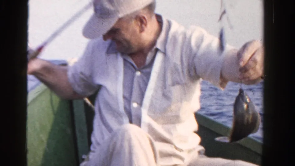 The fisherman sits in the small boat and holds up a line with a small fish, continuing the 1958 Ontario fishing scene.