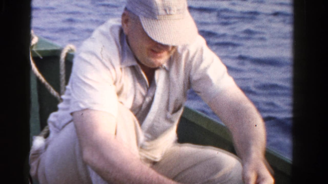 Man in a light shirt and cap leans over fishing gear in a small boat on a lake.
