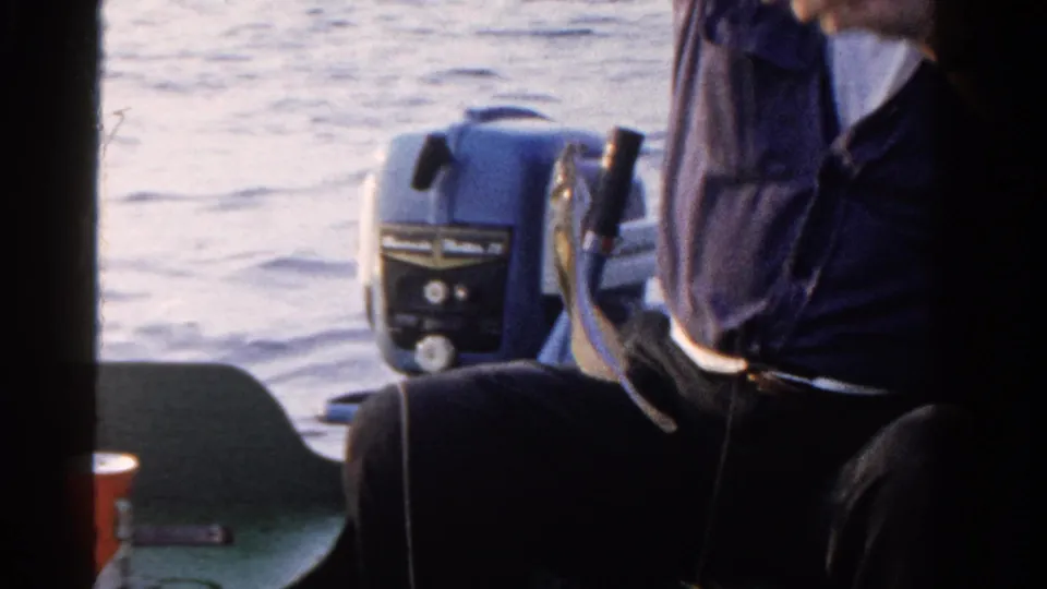 A man in dark clothing sits in the small boat while holding fishing line, with lake water and the outboard motor behind him.