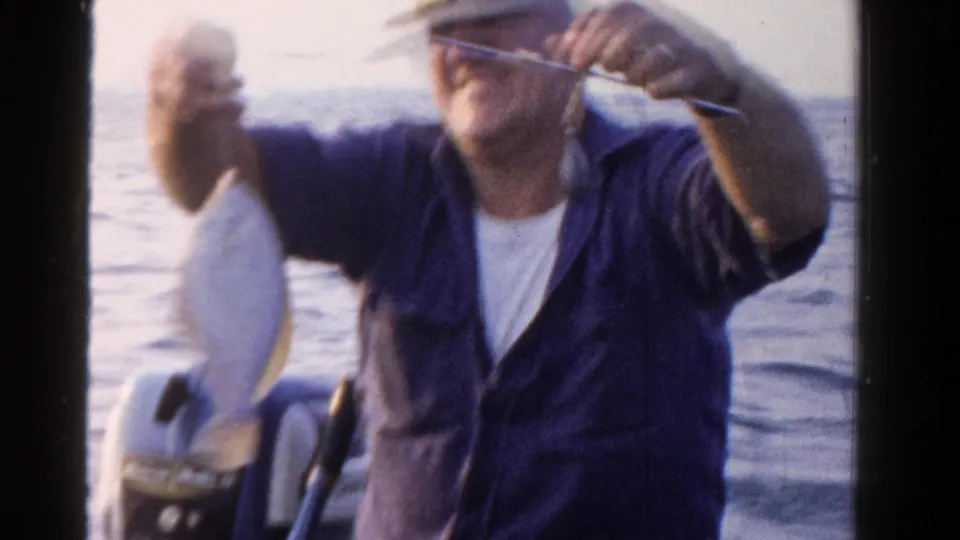 The man lifts the fish and line toward the camera, with open water and the boat motor visible behind him in the Ontario home movie.