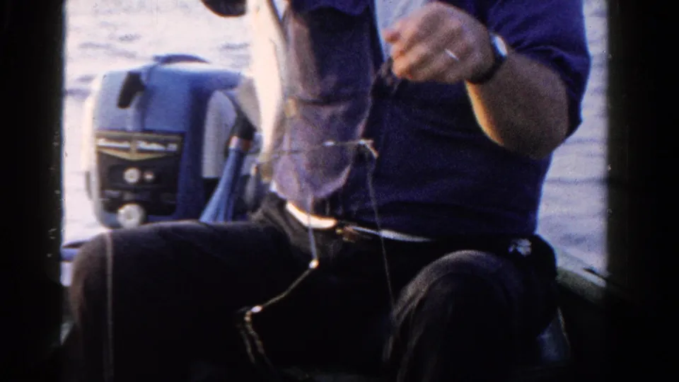 In the boat, the man lowers the catch and works with the fishing line or hook, with the motor and water still visible behind him.