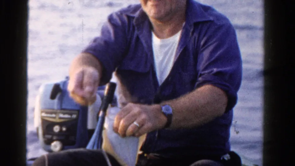 The man sits in the small boat and holds the fish lower near his lap, with open water and the outboard motor in the background.