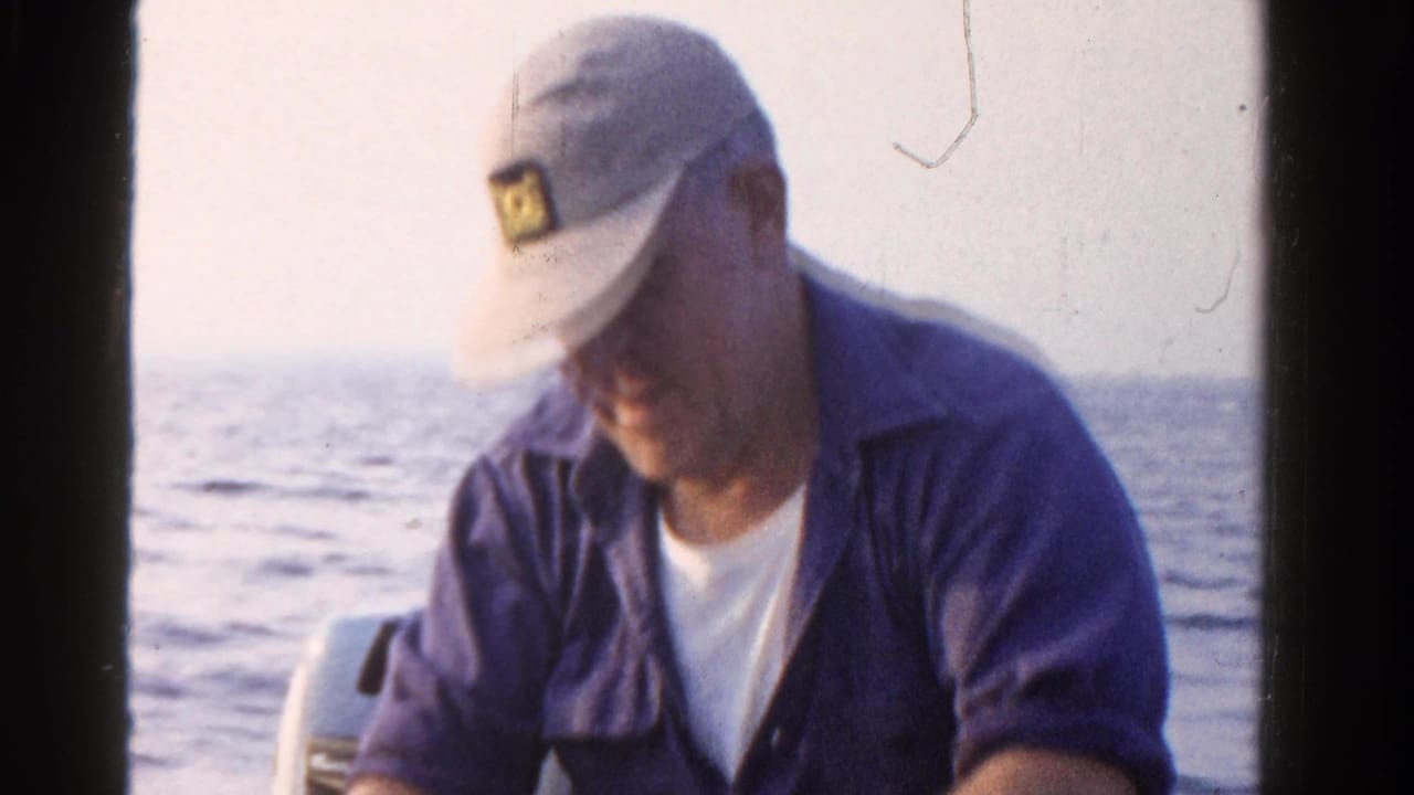 In 1958 Ontario home-movie footage, a seated fisherman in a white cap and blue shirt looks down while working near his catch on a boat.