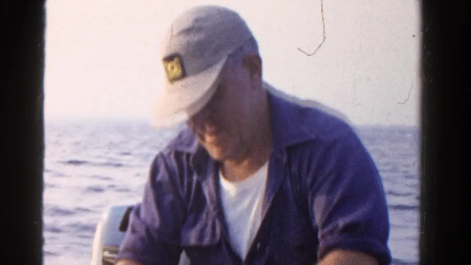 In 1958 Ontario home-movie footage, a seated fisherman in a white cap and blue shirt looks down while working near his catch on a boat.