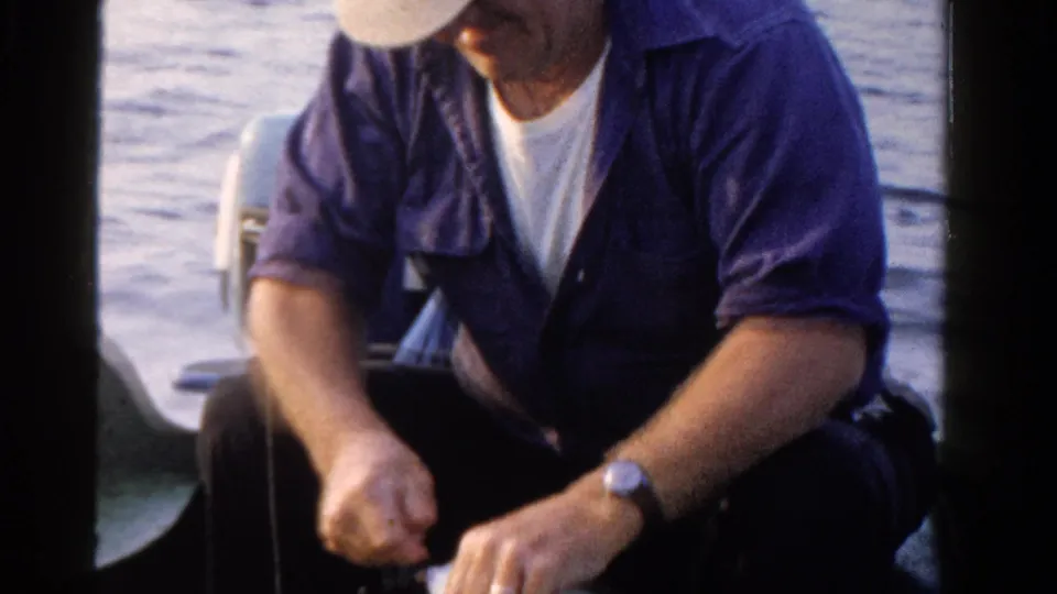 The fisherman sits in the boat with his hands close to the fish or line, with open water and part of the motor visible behind him.