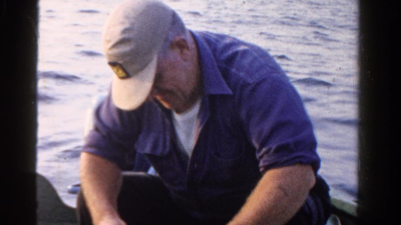 Man in a cap and blue shirt sits in a boat beside calm water while looking down at his hands.