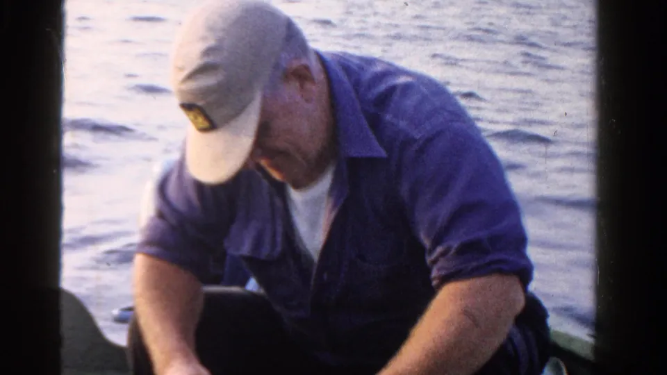 In 1958 Ontario home-movie footage, a man in a light cap and blue shirt sits on a boat and looks down while handling fishing gear.