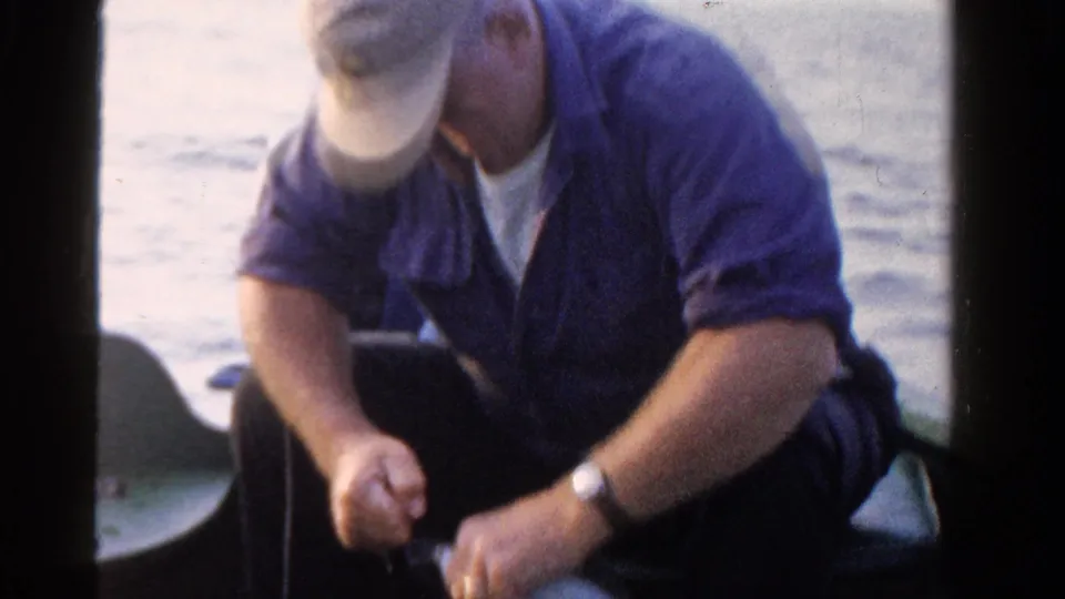 A soft, grainy 8mm frame shows the capped man bending forward in the boat as he continues preparing fishing gear.