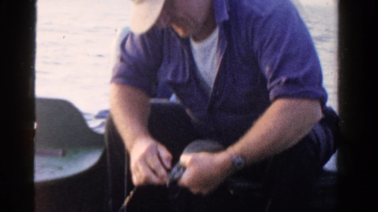In 1958 Ontario home-movie footage, a seated man in a cap handles a reel or line while fishing from a small boat.