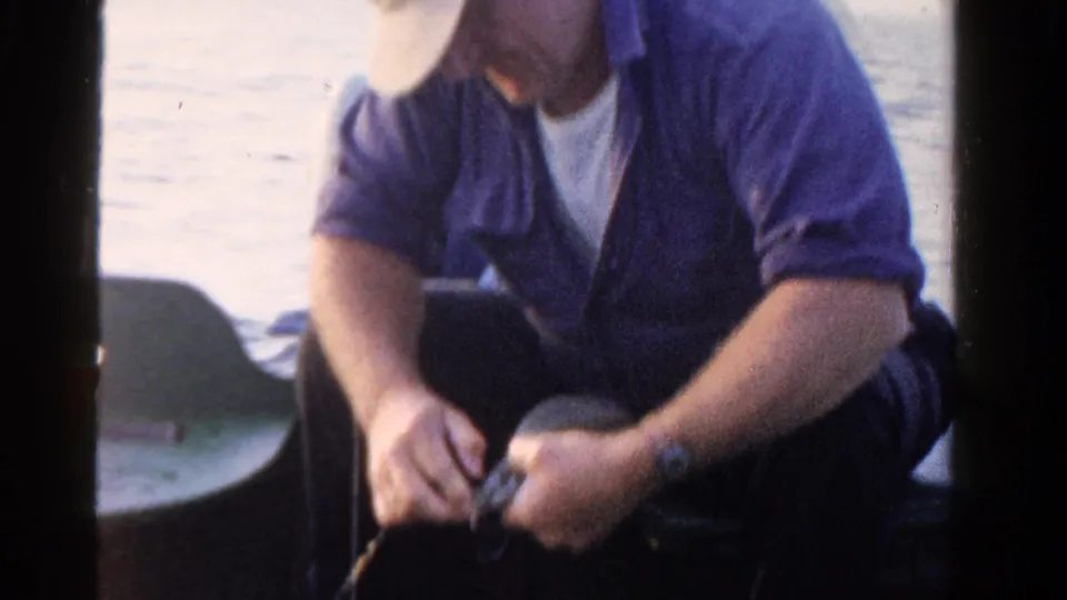 In 1958 Ontario home-movie footage, a seated man in a cap handles a reel or line while fishing from a small boat.