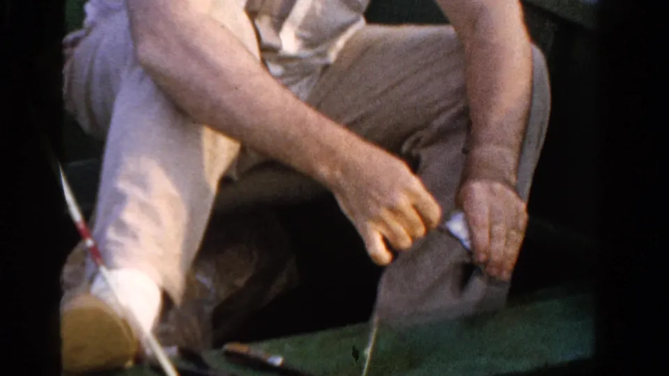 The angler sits in the small boat with fishing line near his hands, light clothing visible in soft 8mm color.