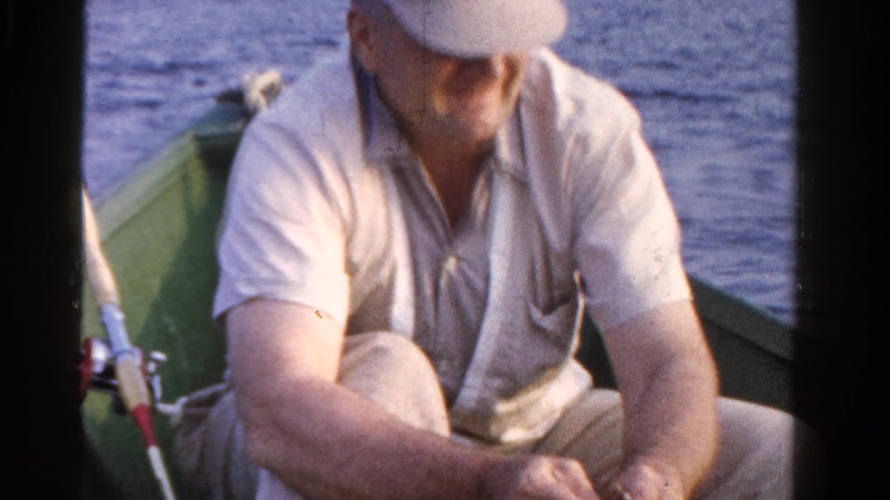Man in a light cap sits in a green boat while handling fishing tackle near lake water.