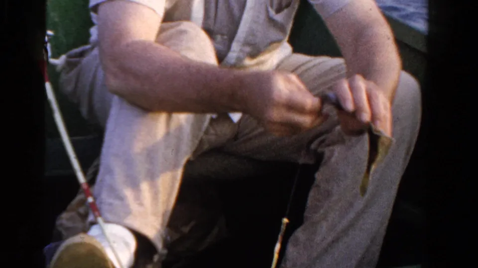 A close 1958 Ontario boat scene shows the seated angler's hands working with bait or tackle beside fishing rods and lake water.