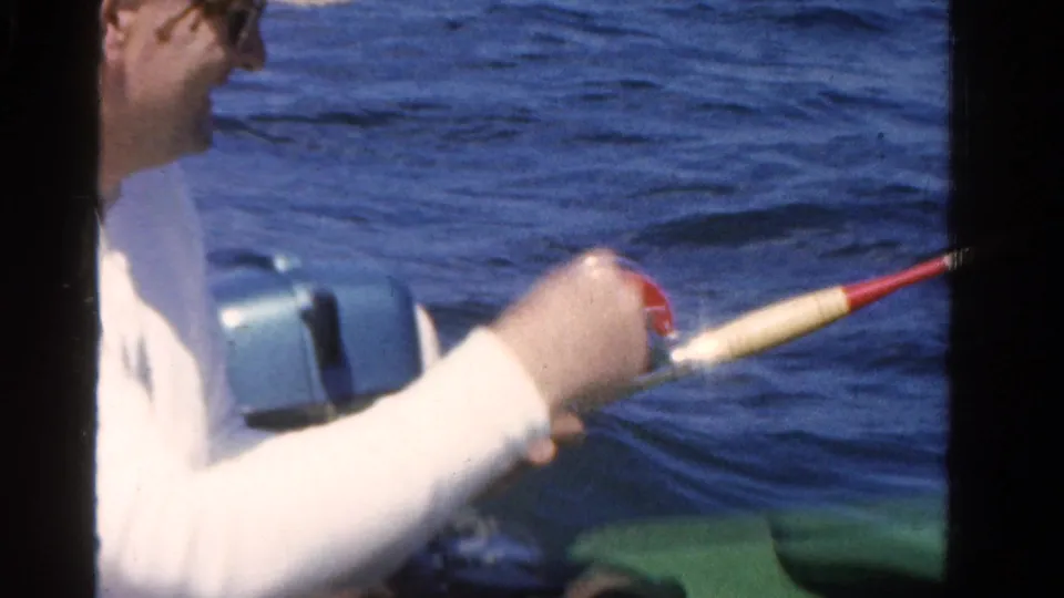 A person in a white shirt handles a fishing rod from the green boat, with blue lake water and an outboard motor visible nearby.