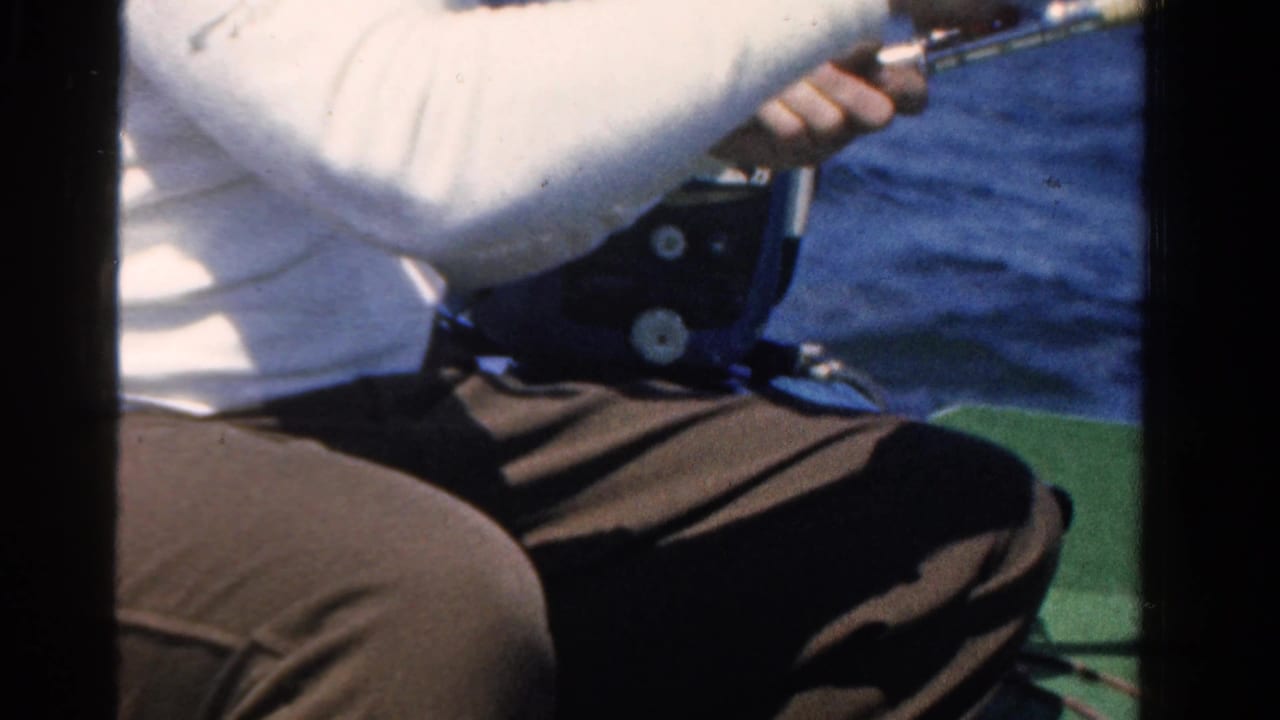 Seated person in a small boat holds a fishing rod over blue water.