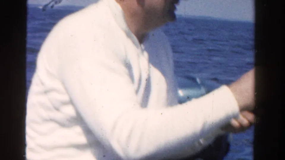 Middle-aged man in a white shirt reels a fishing line from a boat on Ontario water in the 1958 home movie.