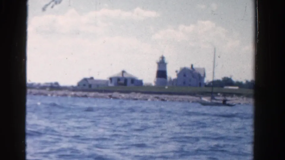 Ontario shoreline seen from the boat, with low buildings, a lighthouse-like structure, blue water, and a small sailboat nearby.