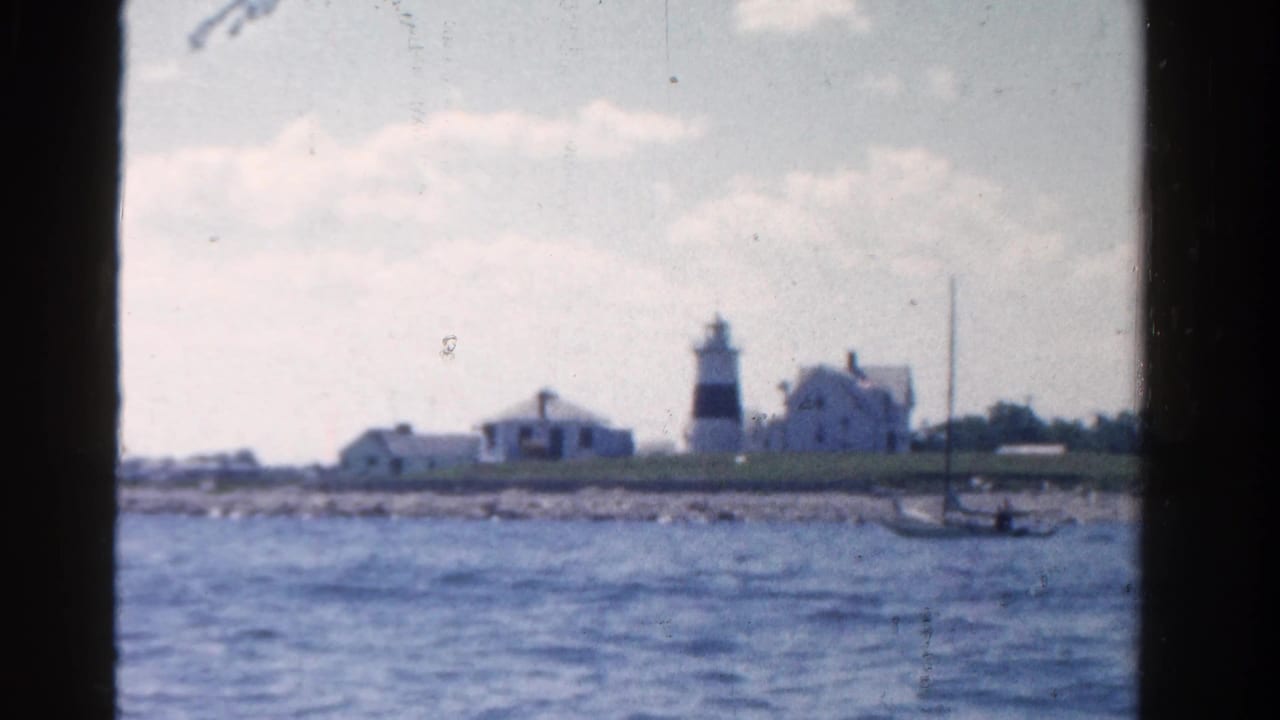 Distant lighthouse and shore buildings seen across choppy blue water from a boat.