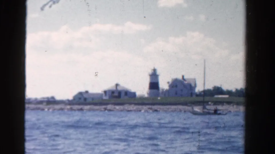 1958 Ontario 8mm frame showing a lighthouse and white shoreline buildings across rippling lake water from a moving boat.