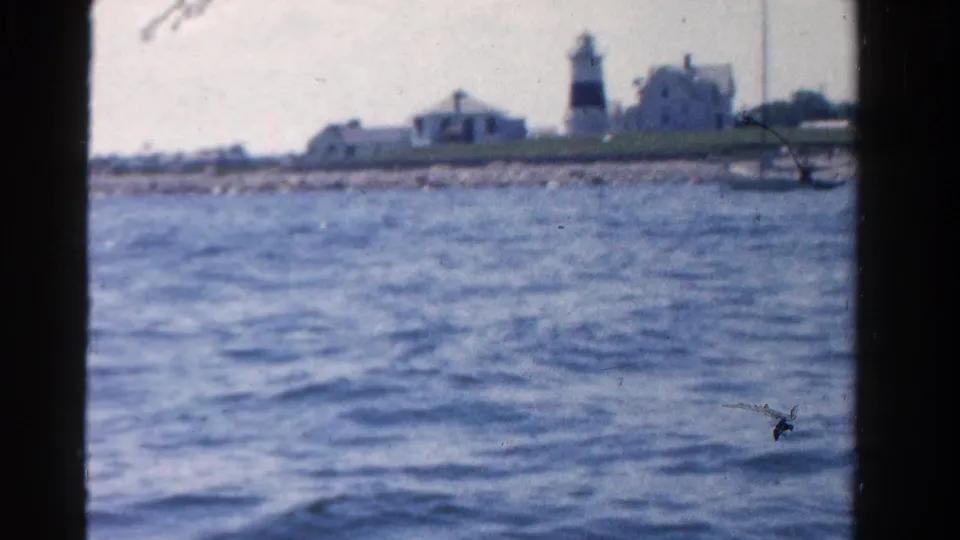 1958 Ontario 8mm view of a lighthouse complex and low shoreline buildings across a broad stretch of blue water.