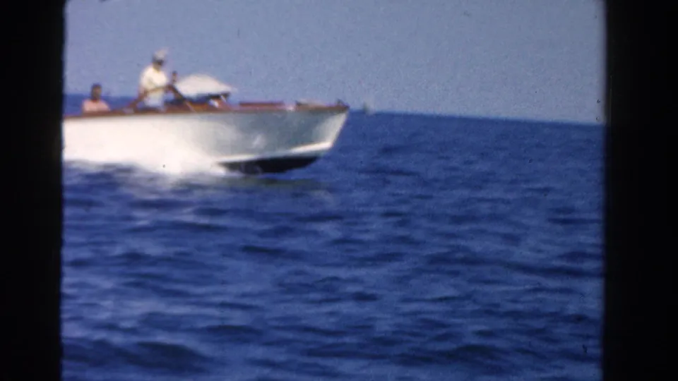 A closer 1958 Ontario home-movie frame catches the white speedboat partly cropped as it passes, with passengers just visible.