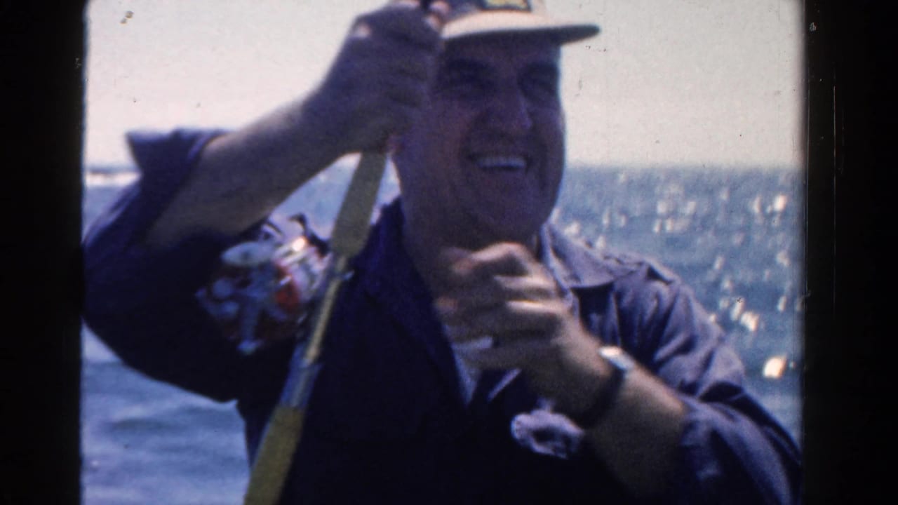 Smiling man in a cap holds a fishing rod and reel on a small boat with water behind him.
