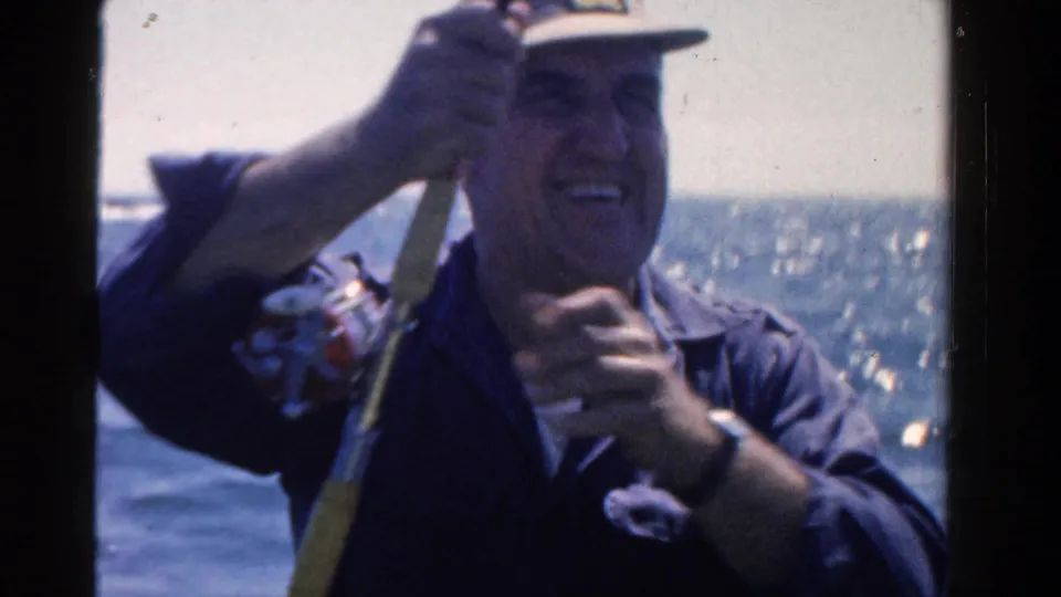 In 1958 Ontario home-movie footage, a smiling man in a cap holds a fishing rod and reel aboard a small boat on open water.