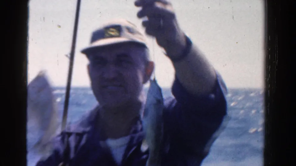 A man in 1958 Ontario home-movie footage raises his fishing line as a caught fish hangs beside him on a small boat.
